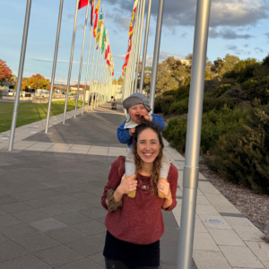 Anna carrying Leo on her shoulders standing in front of a row of flags poles